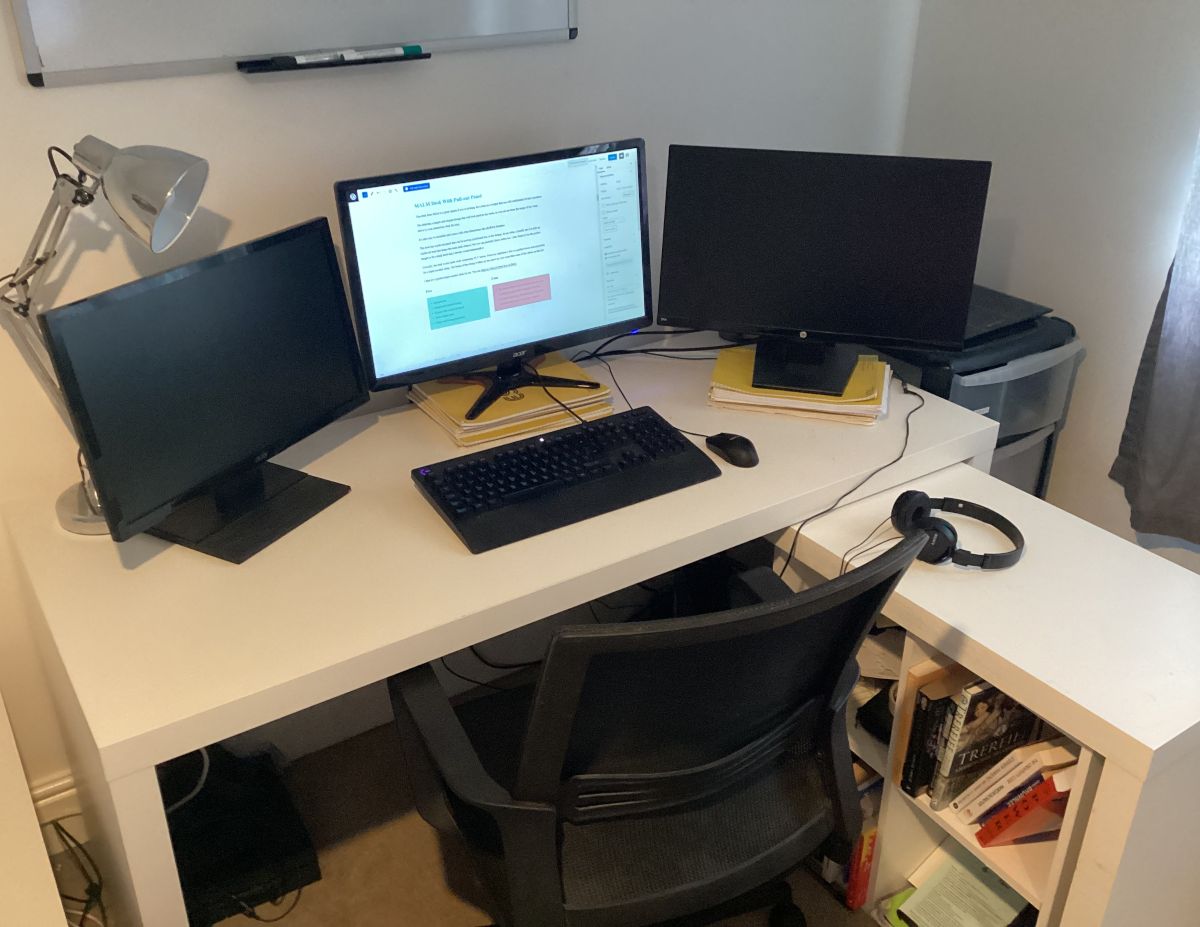 My MALM desk with pullout side table. The desk has 3 monitors, a lamp, a keyboard, a mouse and a pair of headphones on it. It has a small bookshelf under the pullout.